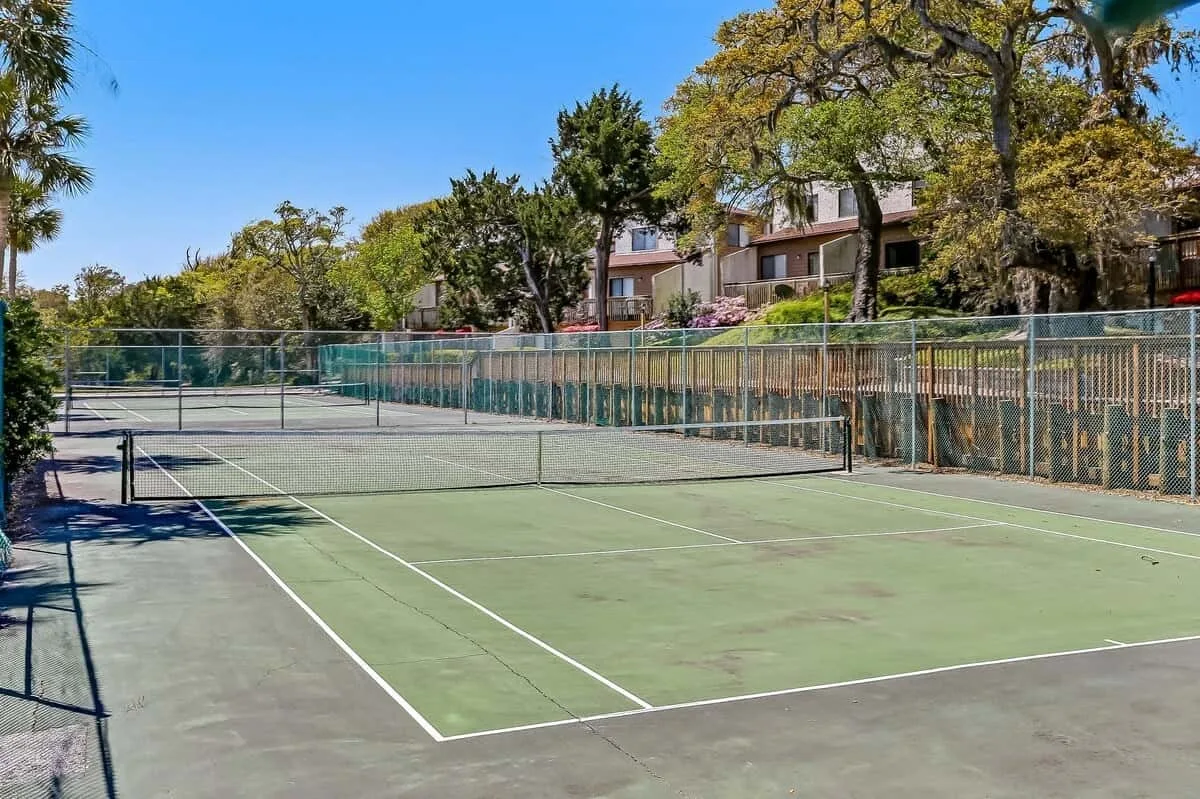 An outdoor tennis court is enclosed by fencing, with trees and buildings beyond.