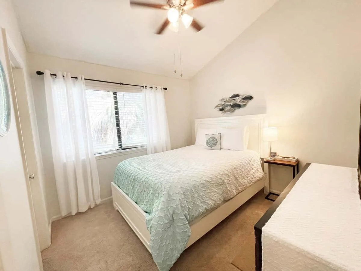 A bright bedroom with a bed beside a curtained window and a ceiling fan.