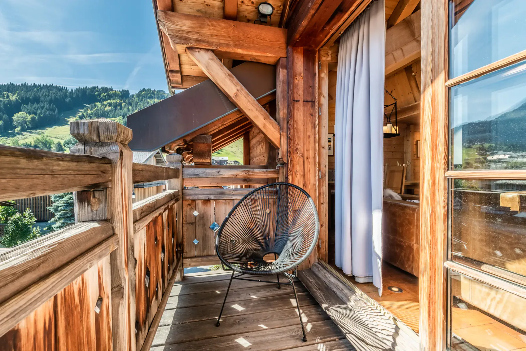 A wooden balcony with a woven seating chair and forested hills in the distance.