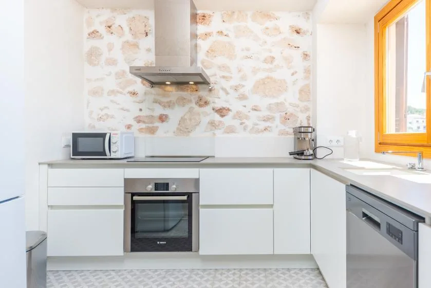 Una cocina luminosa con gabinetes blancos, horno y salpicadero de aspecto pétreo.