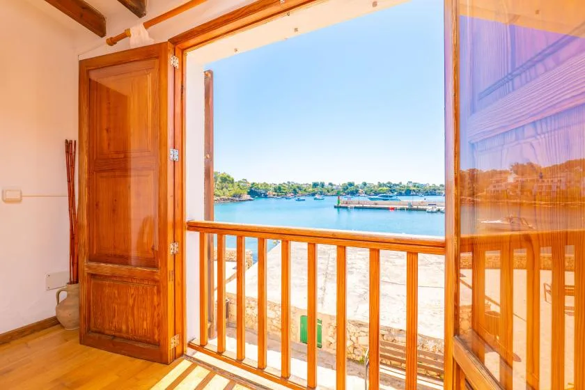 Una puerta de madera abierta da a una vista costera con barcos y agua azul.