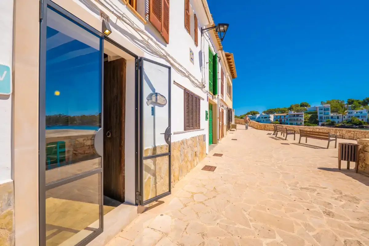 Un paseo soleado y pavimentado bordea edificios blancos con puertas de vidrio abiertas y asientos al aire libre.