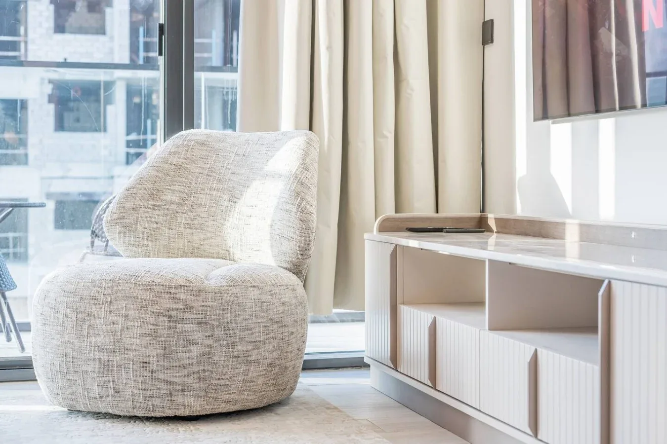 Una sala luminosa con un sillón tapizado claro junto a una consola baja y ventanales.