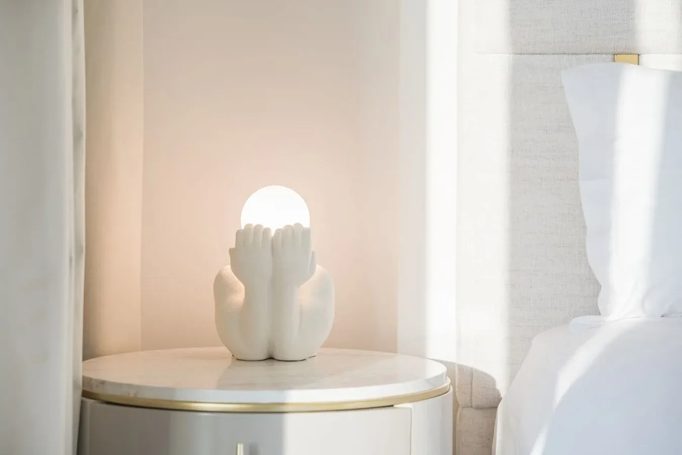 Una esquina de dormitorio suavemente iluminada presenta una lámpara escultórica sobre una mesa redonda de noche.