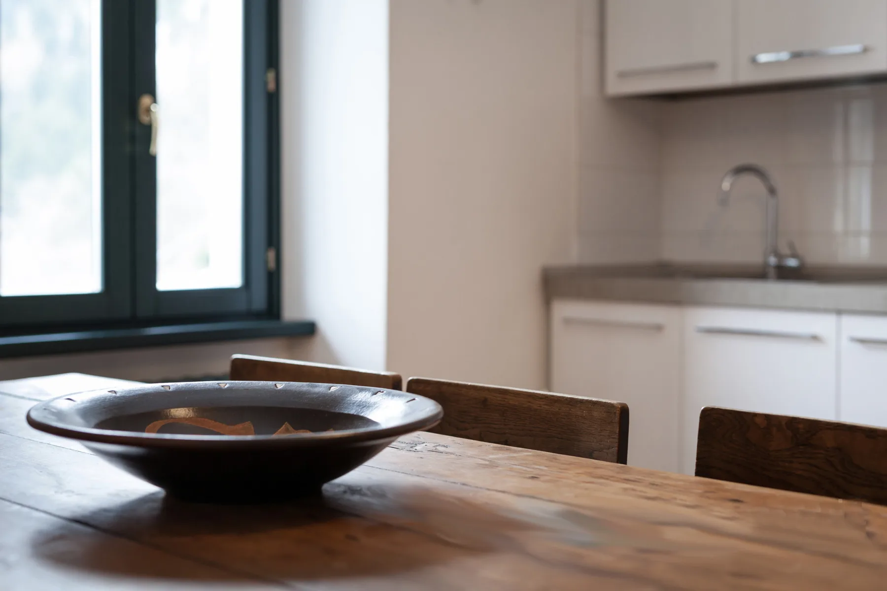 A wooden dining table with a shallow bowl sits beside a bright kitchen.