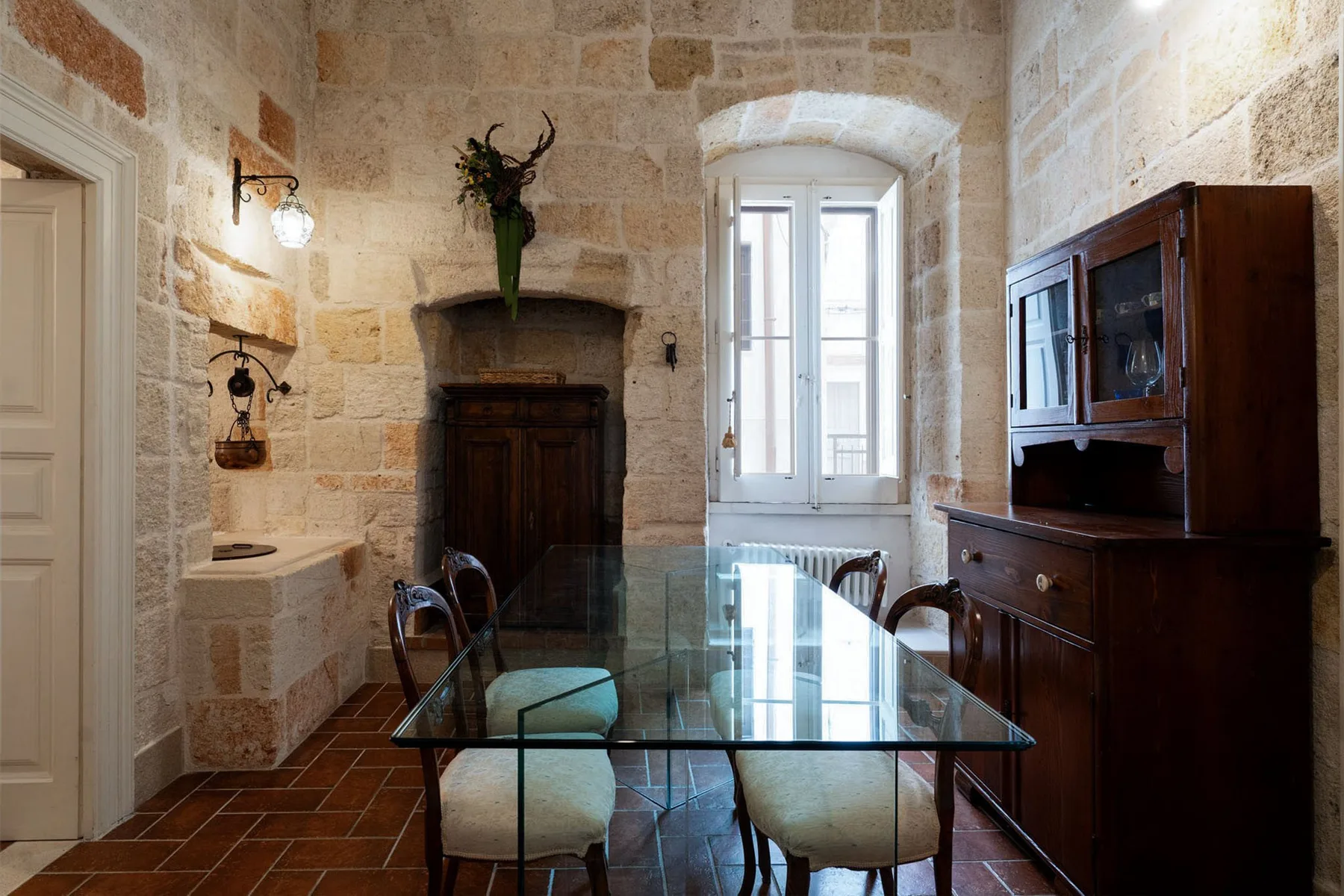 A stone dining room with a glass table and dark wooden cabinet.