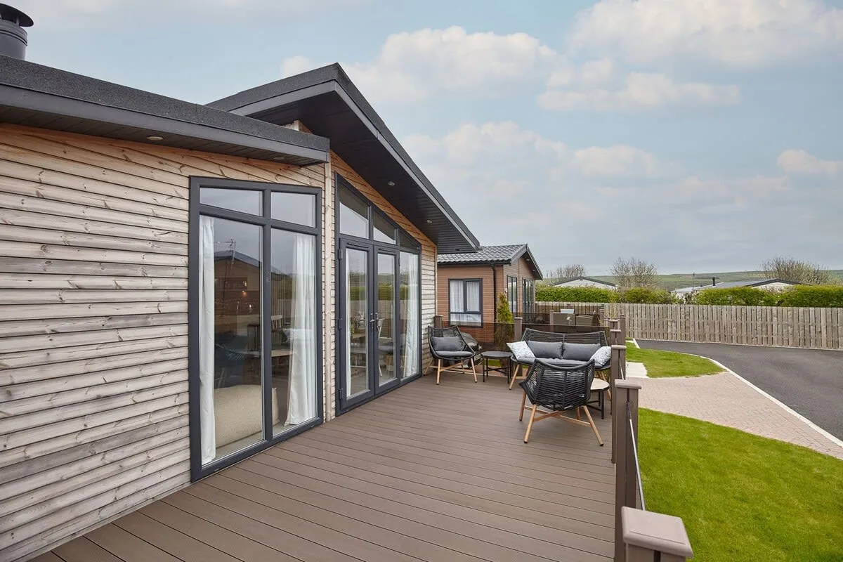 A modern home opens onto a wooden deck with outdoor seating and a lawn.
