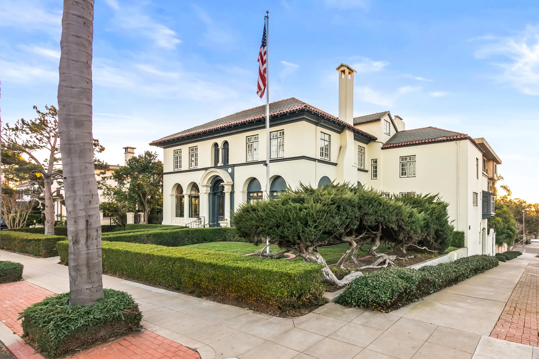 A large cream-colored building with arched windows stands behind a landscaped hedge.