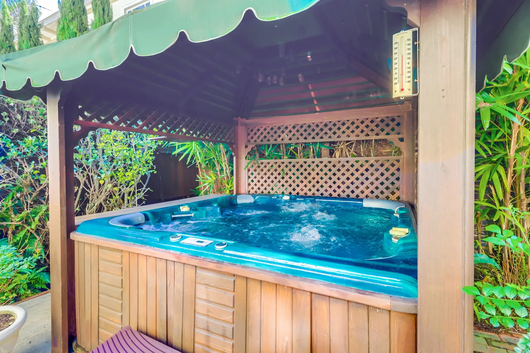 A bubbling hot tub sits beneath a covered wooden pavilion surrounded by greenery.