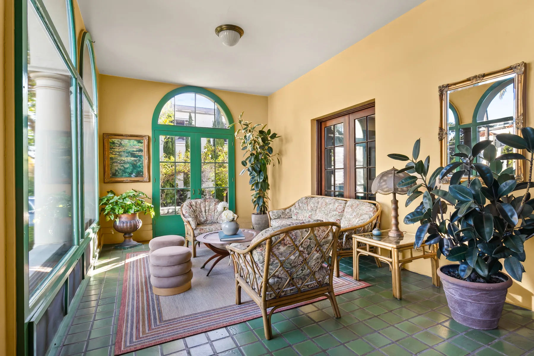 A sunlit sitting room with wicker seating, tall green-framed windows, and potted plants.