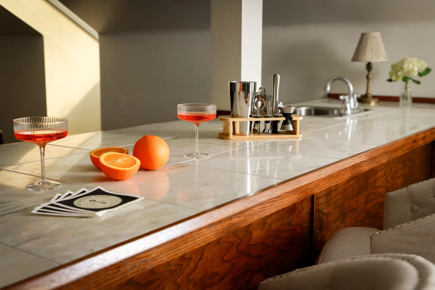 A bright kitchen counter holds two drinks, sliced citrus, and a small flower arrangement.