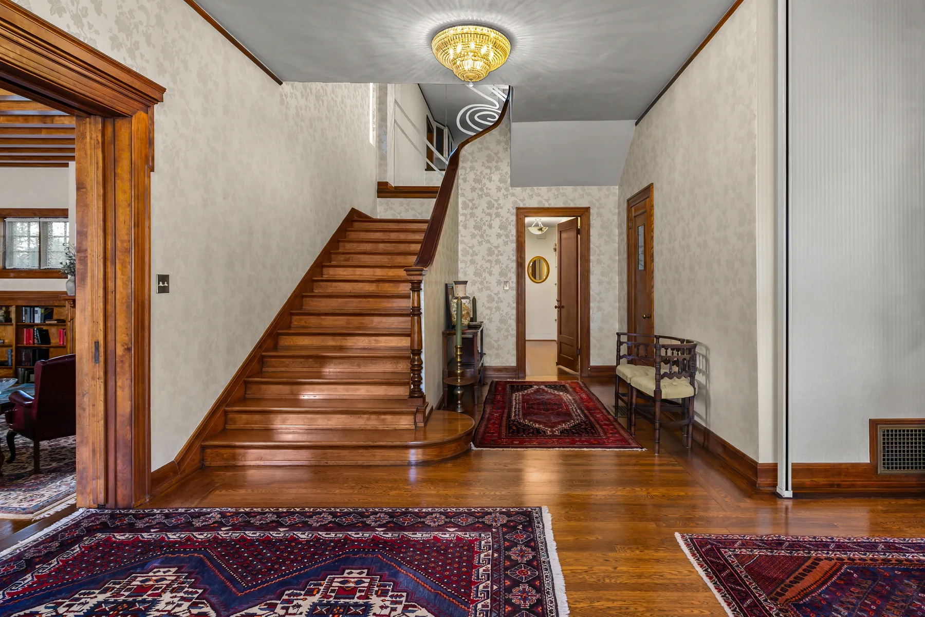 A wood staircase rises through a spacious entry hall with patterned rugs.