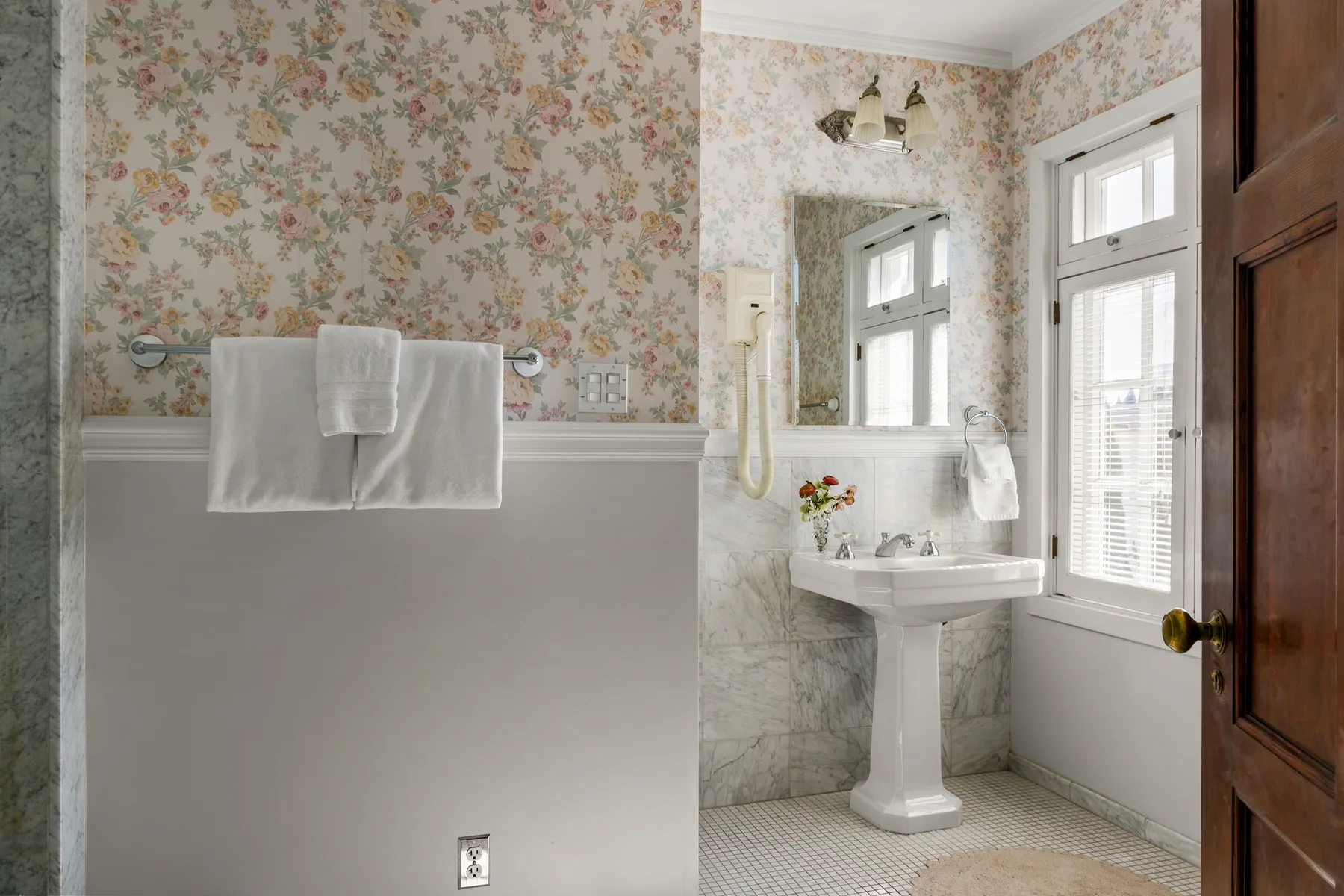 A bathroom with a pedestal sink, mirror, and floral wallpaper.