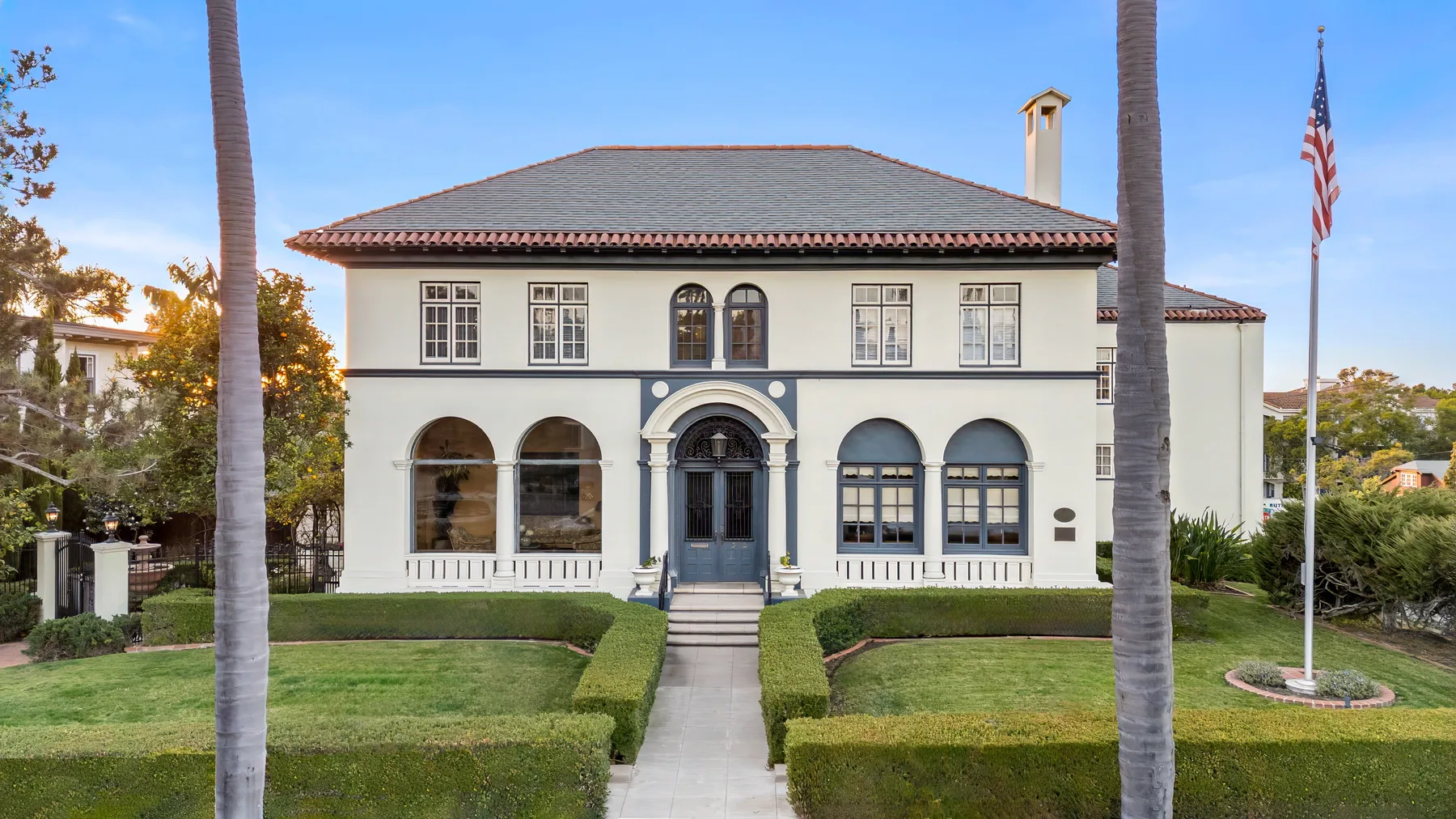 A large two-story building stands behind a straight walkway bordered by trimmed hedges.