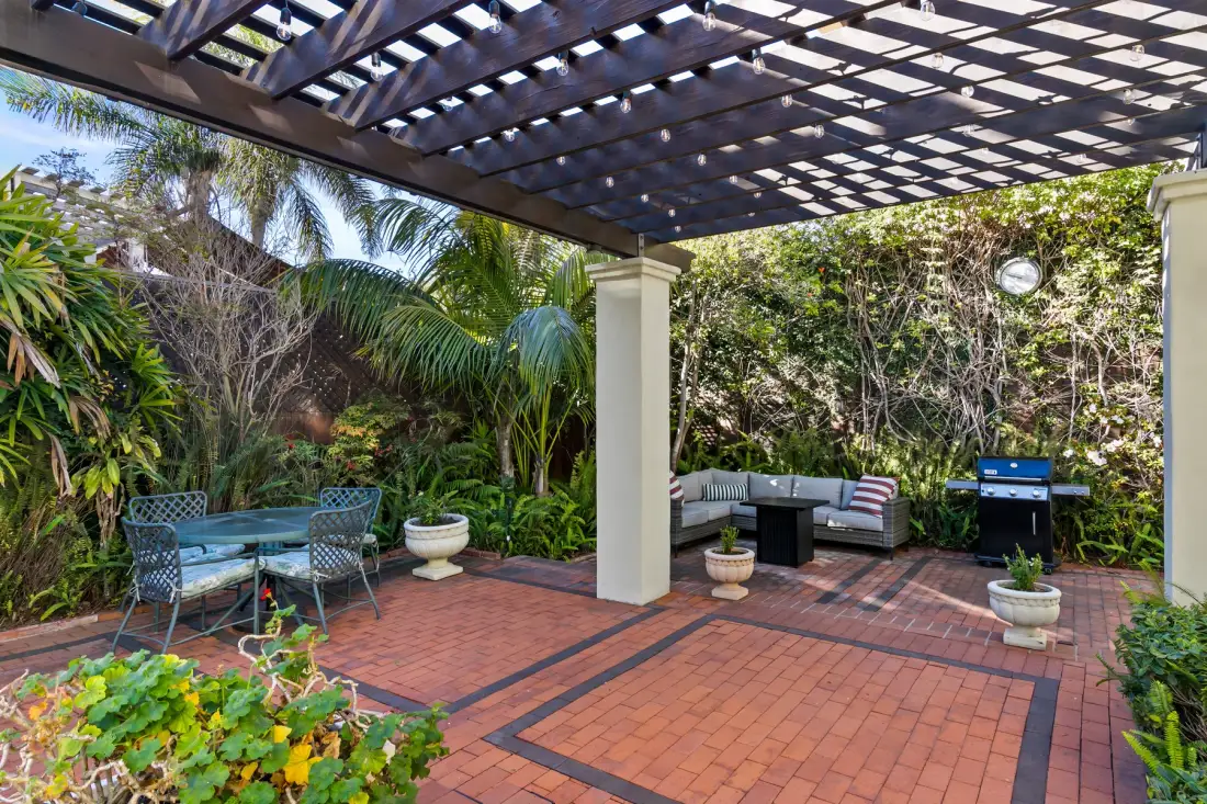 A brick-paved patio under a wooden pergola, with outdoor seating and a grill.