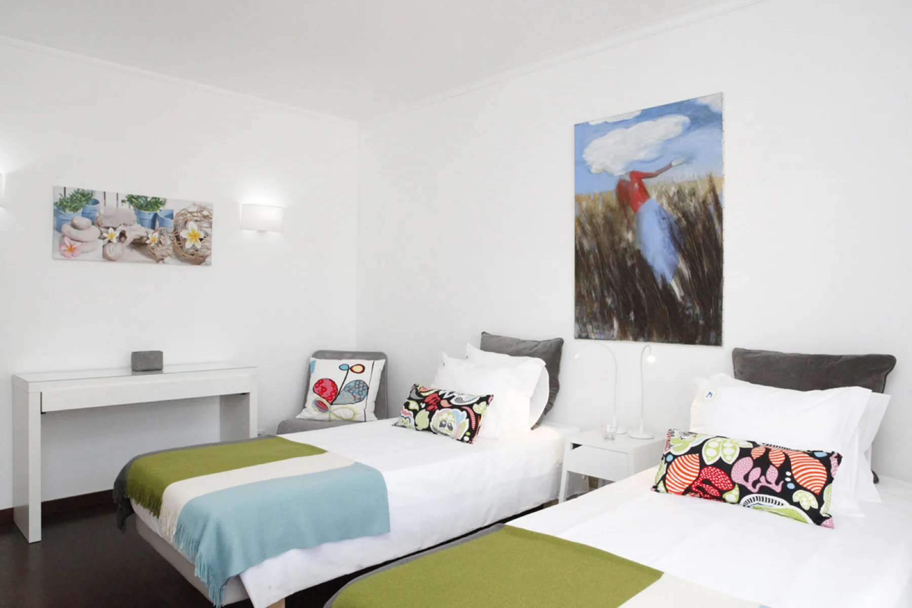 A bright bedroom with two beds, colorful cushions, and wall art.