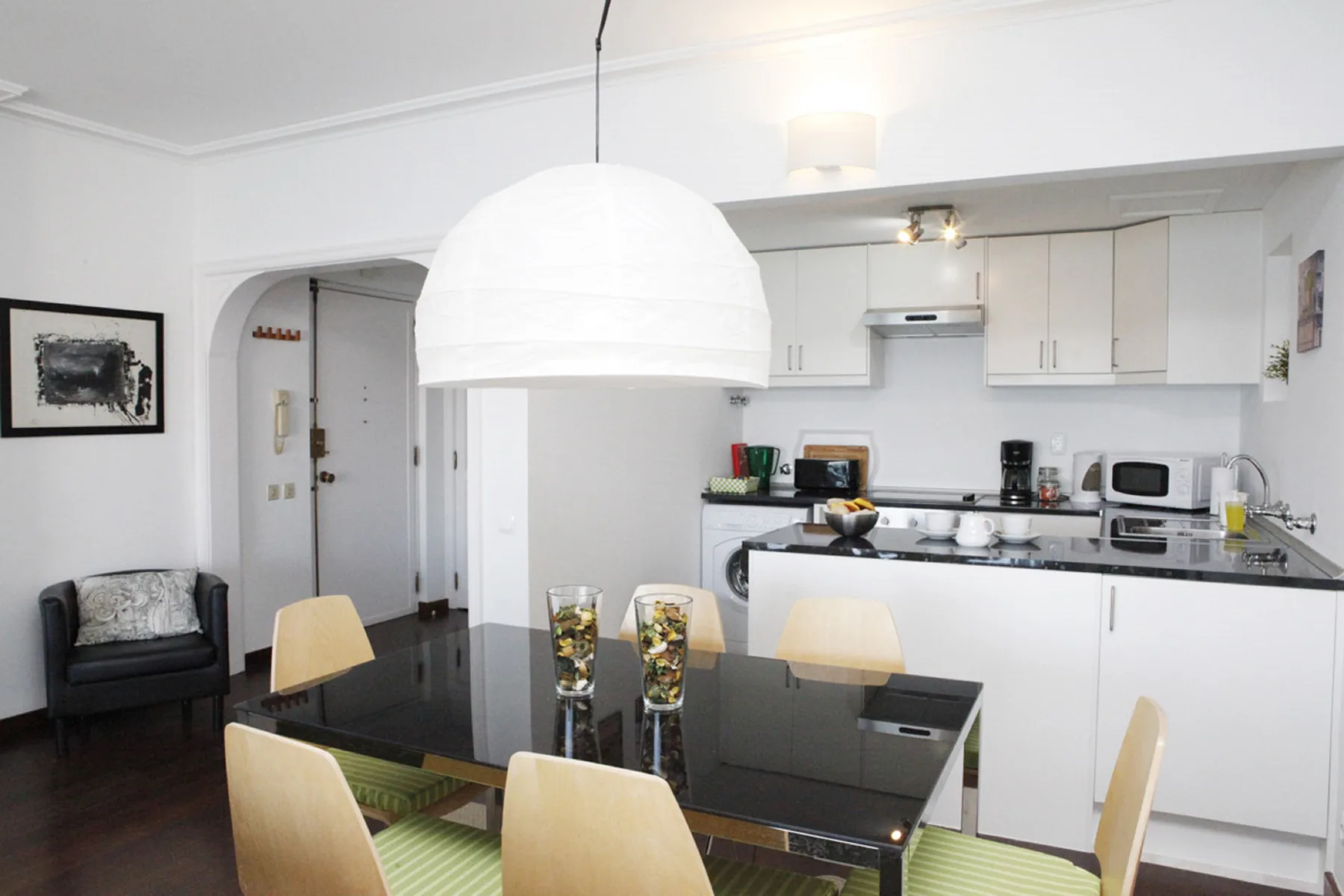 A dining area with a glass table sits beside a white kitchen.