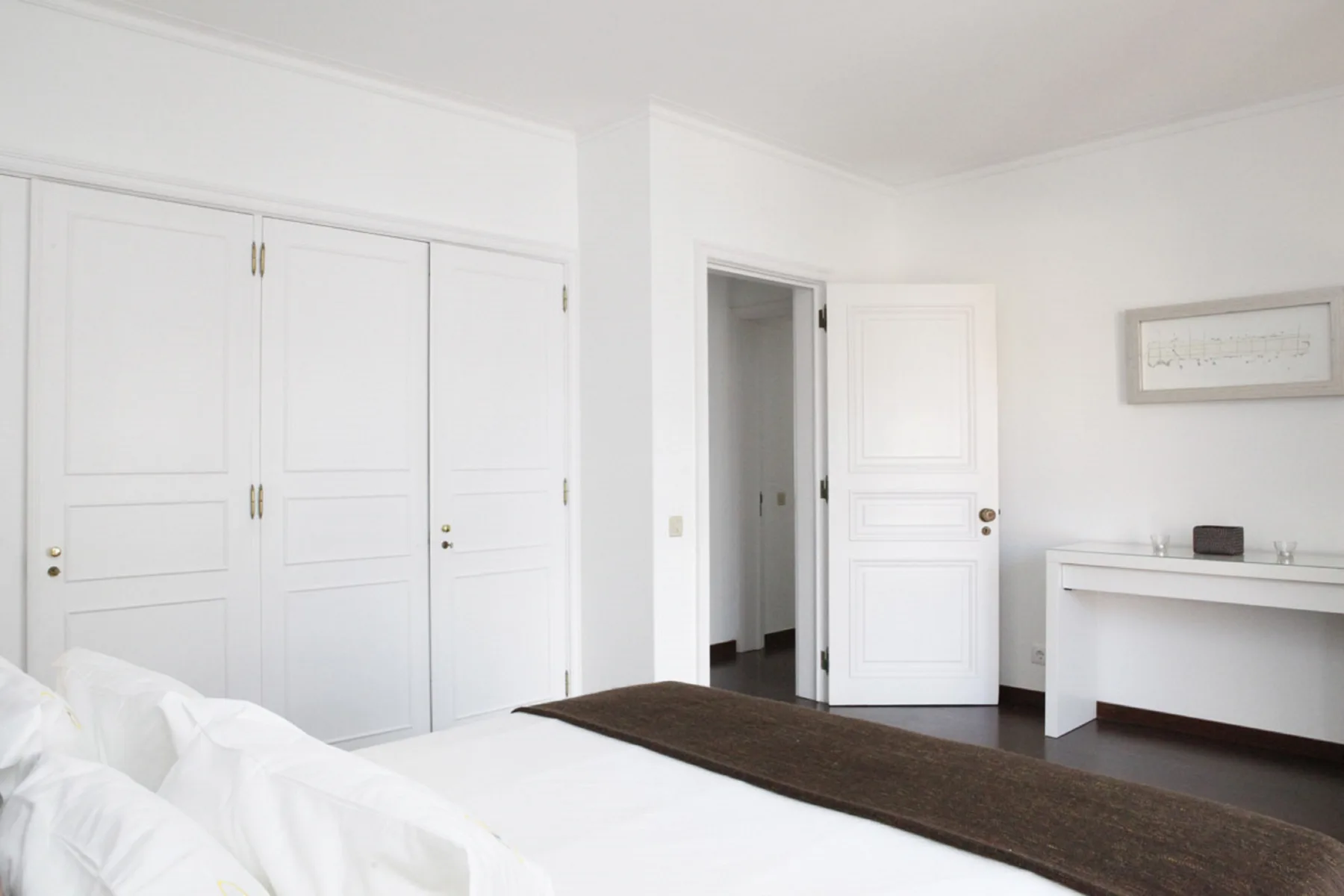 A bright white bedroom with a bed, built-in cupboards, and an open doorway.