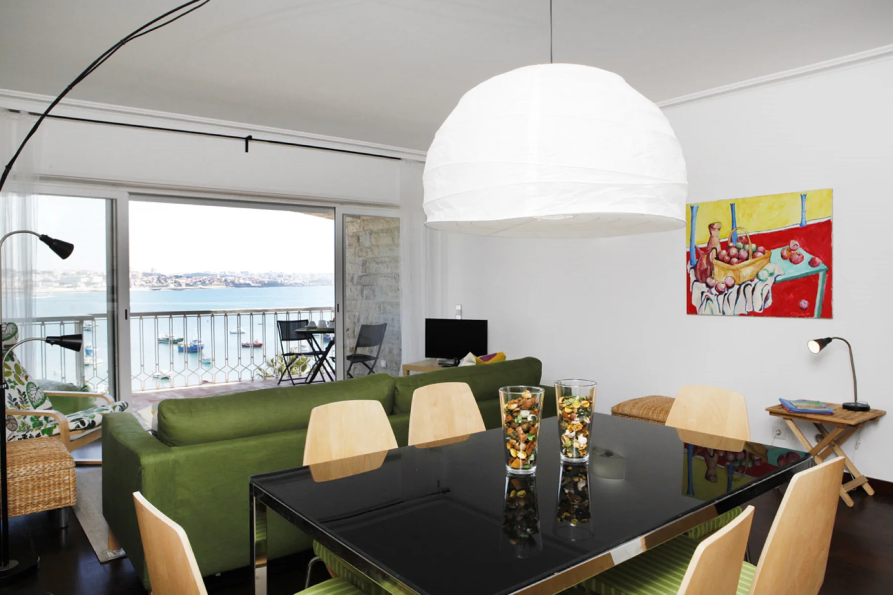 A living and dining area with green seating and a large table beside windows overlooking the water.