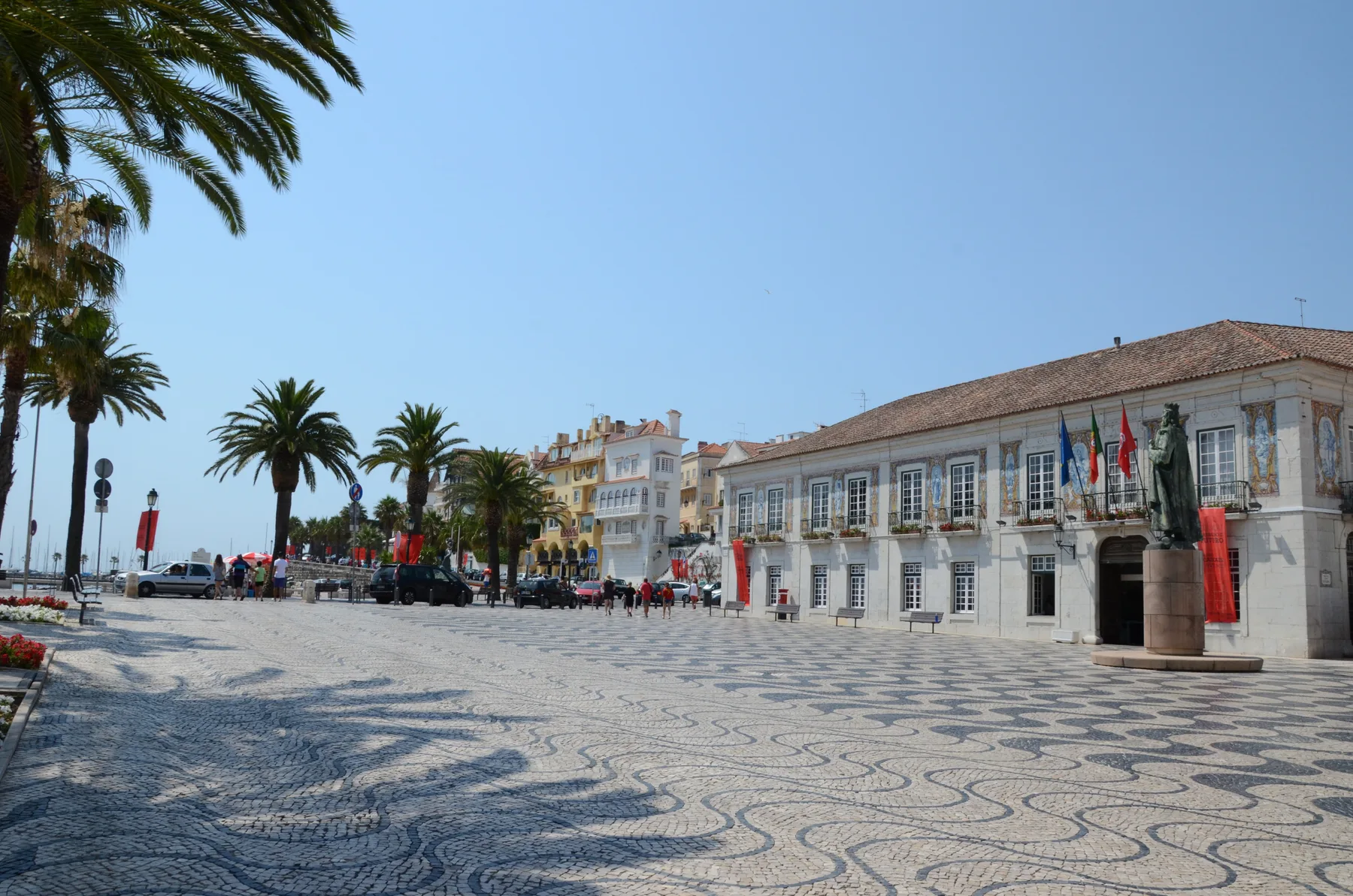 A paved waterfront square with palm trees and a row of buildings.
