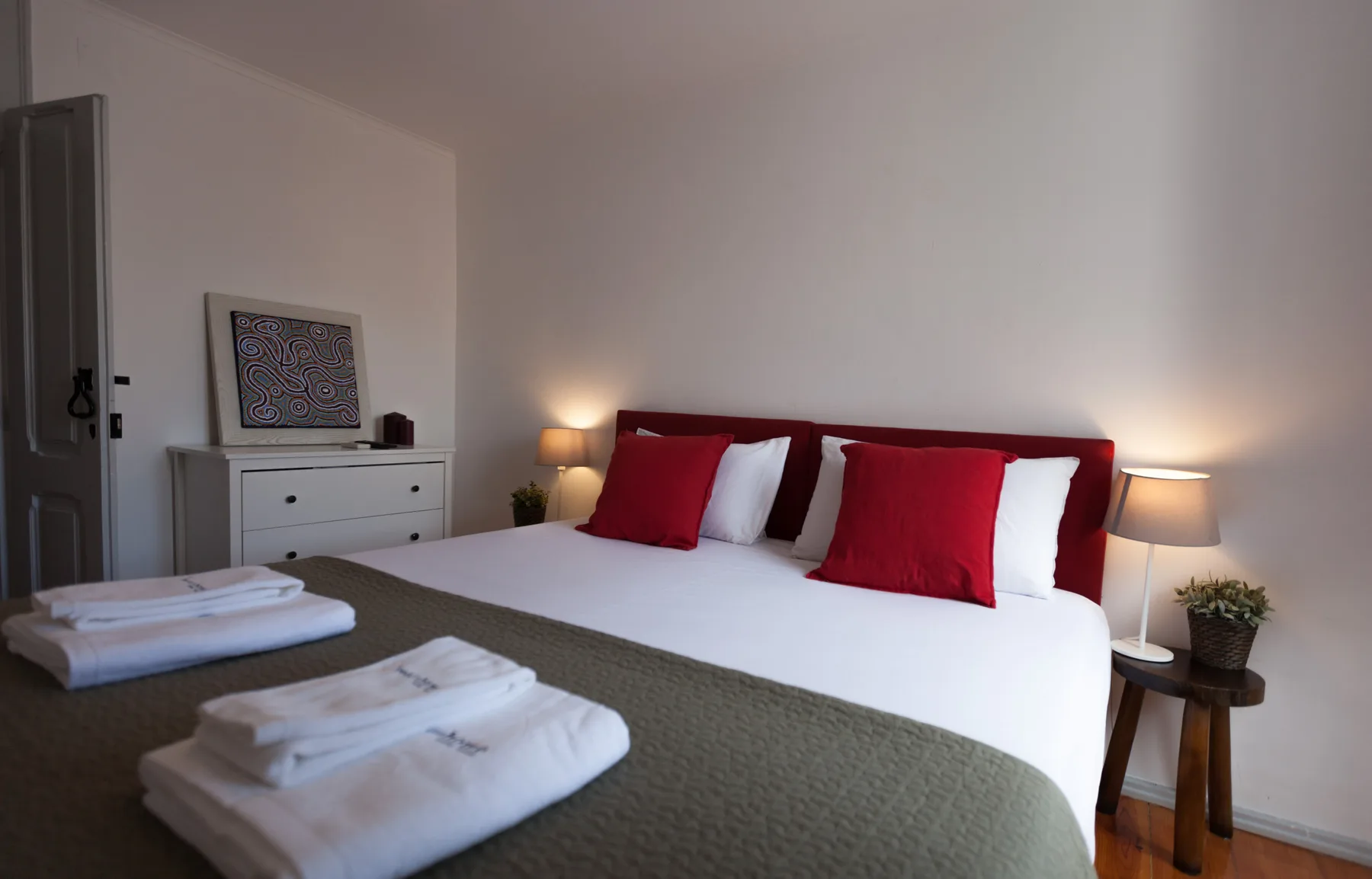 A bedroom with a neatly made bed, red pillows, and bedside lamps.