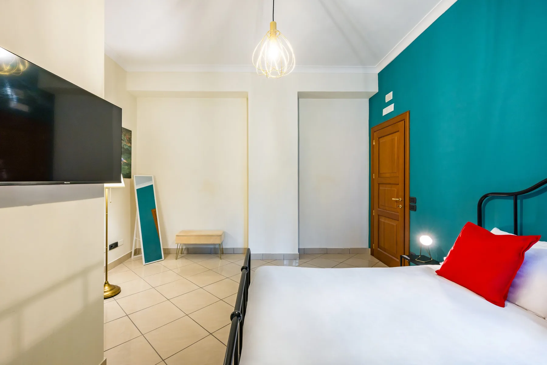 Un dormitorio con una cama blanca, un cojín rojo, un televisor en la pared y una pared turquesa.