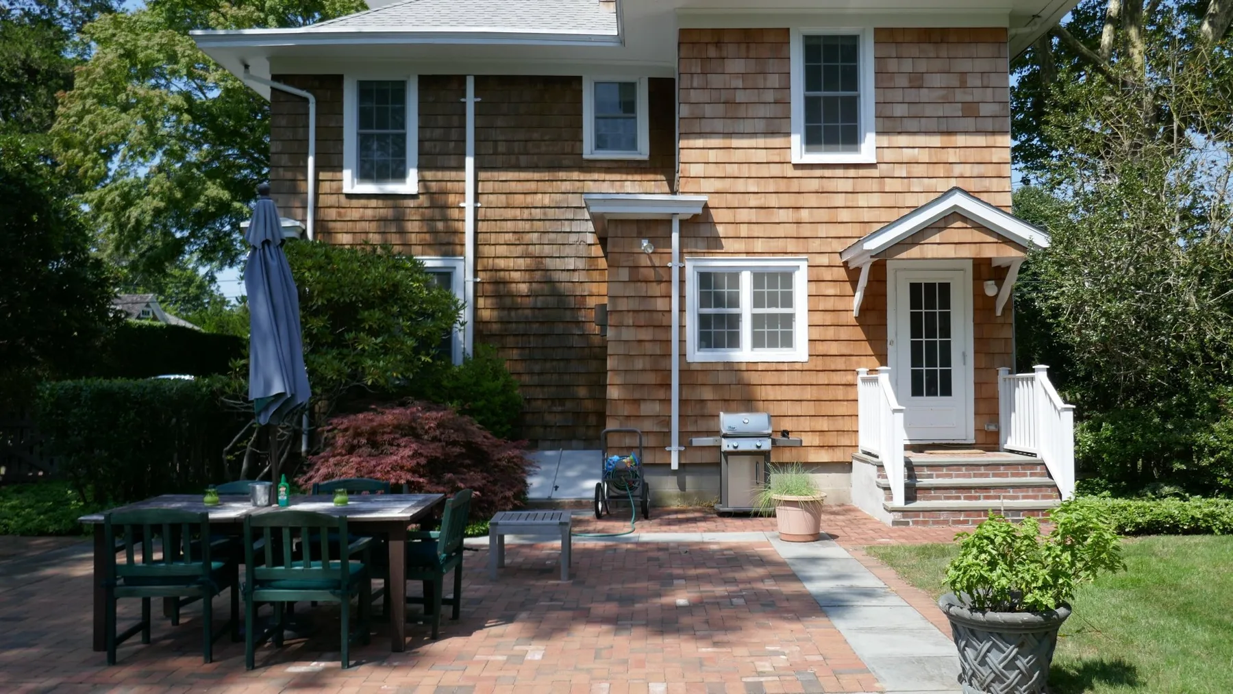 A shingled house stands beside a brick patio with outdoor seating.