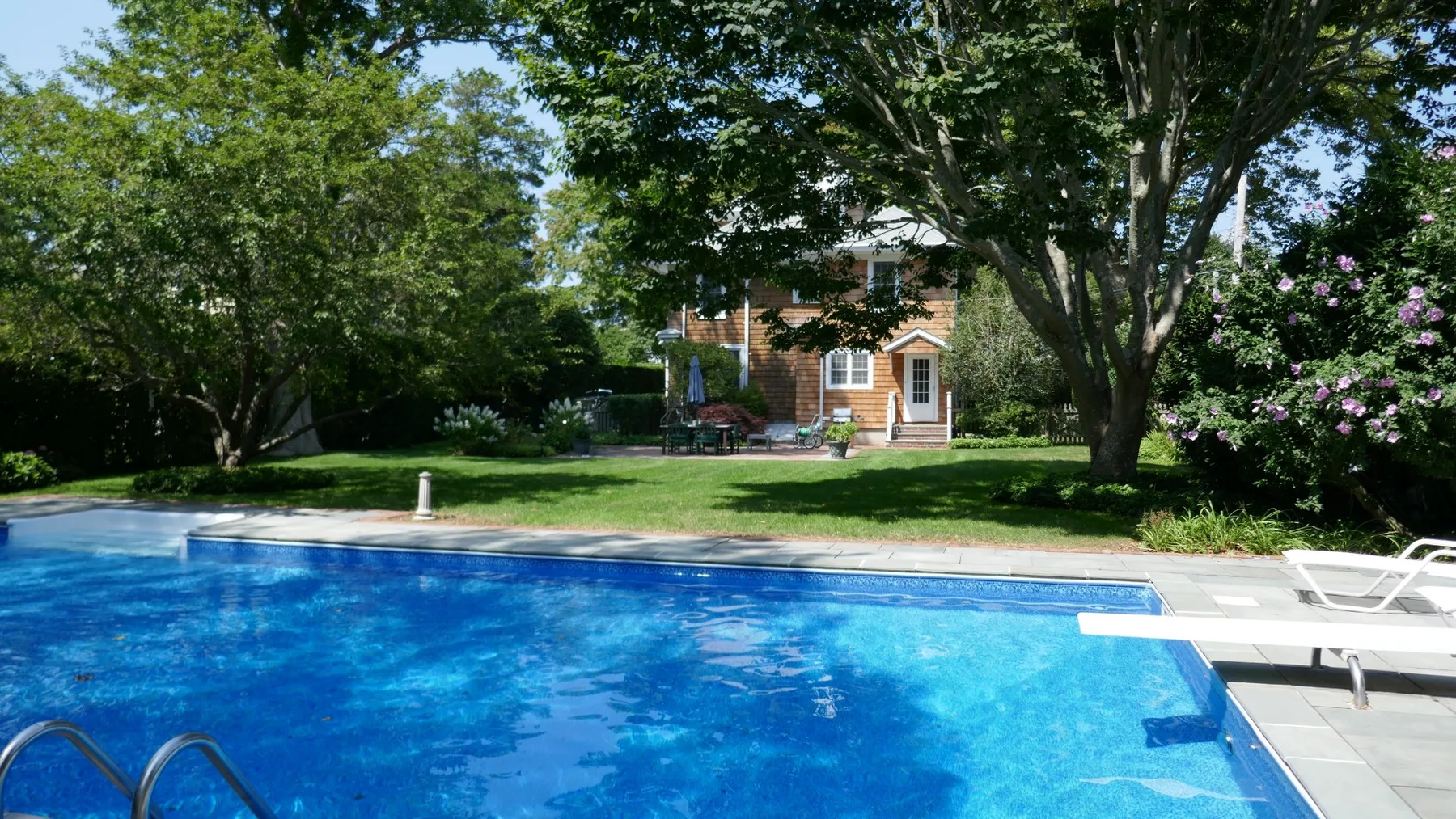 An outdoor swimming pool sits in a landscaped garden with trees and a house beyond.