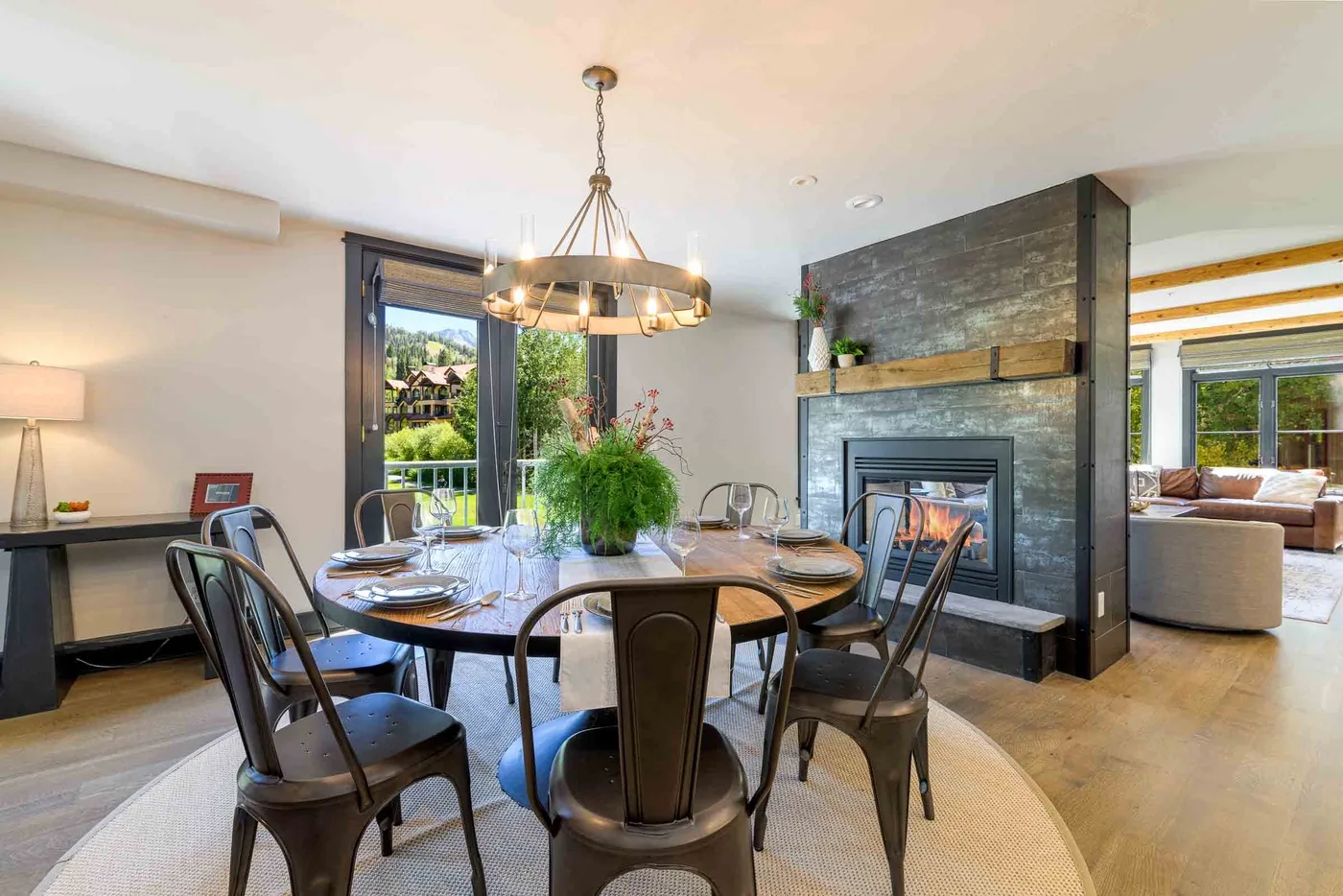 A dining area with a round table beside a stone fireplace and large windows.