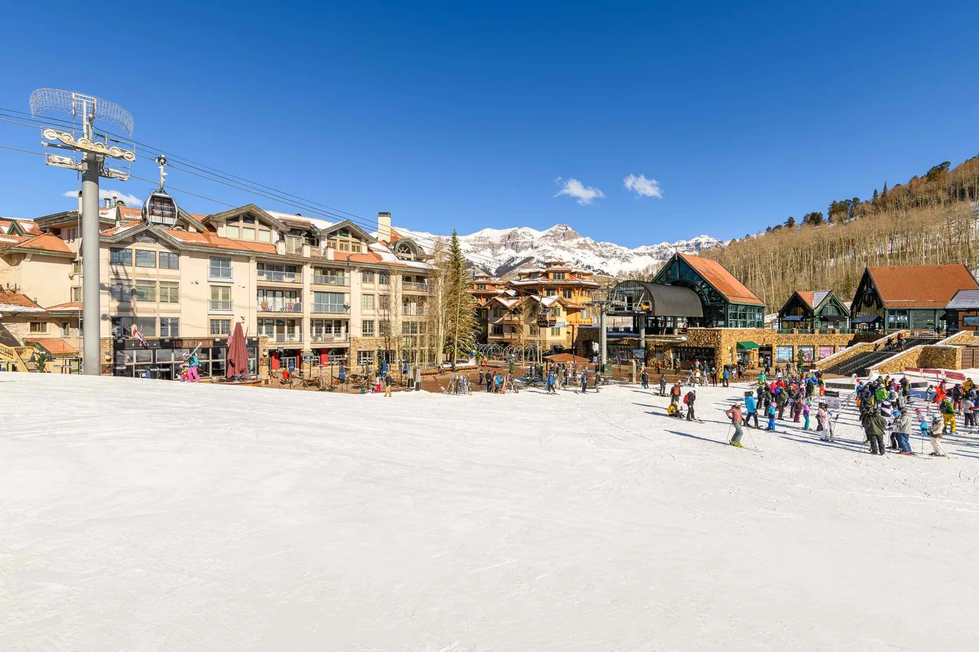 A snowy ski area with people gathered near lodge buildings beneath mountain peaks.