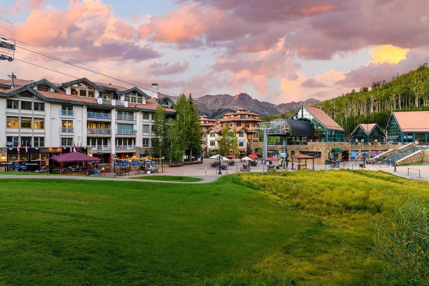 A mountain resort complex beside a grassy slope under a colorful sunset sky.