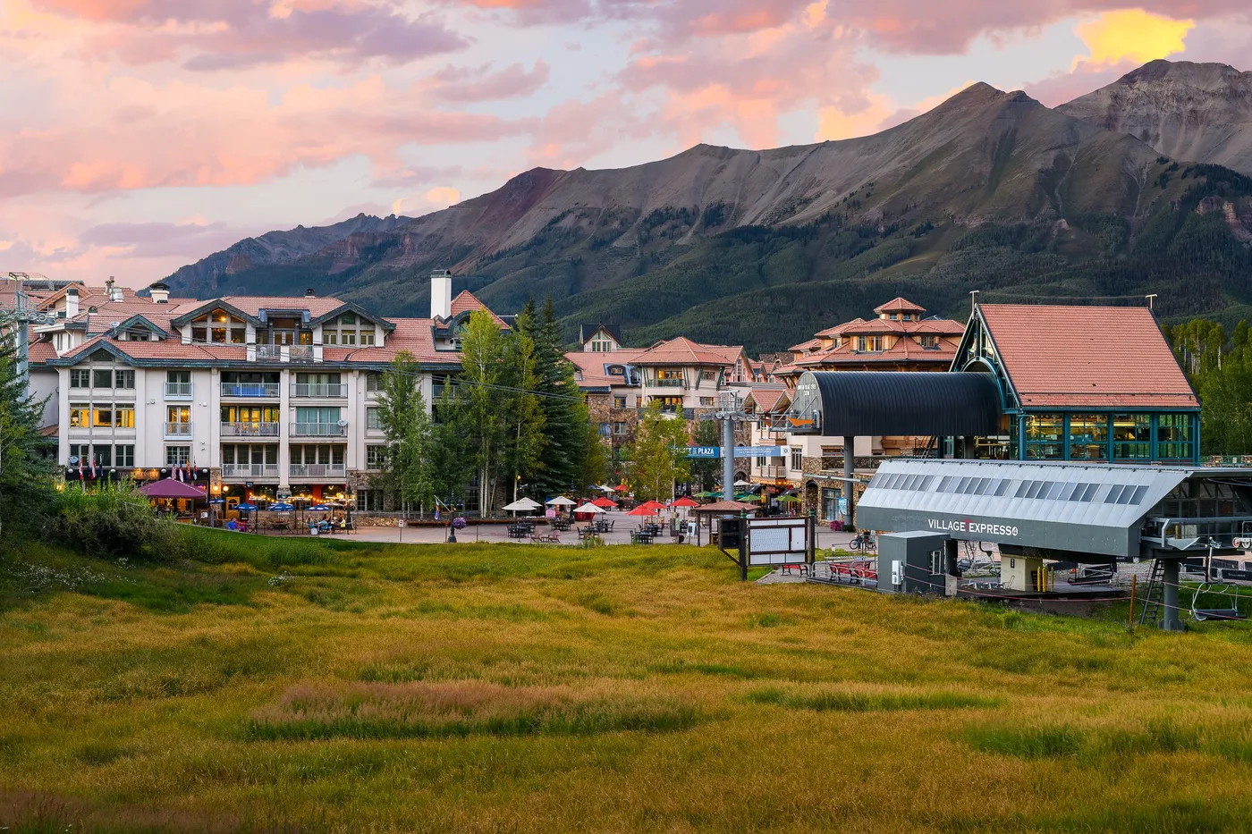 A mountain village with buildings along a grassy open area at sunset.