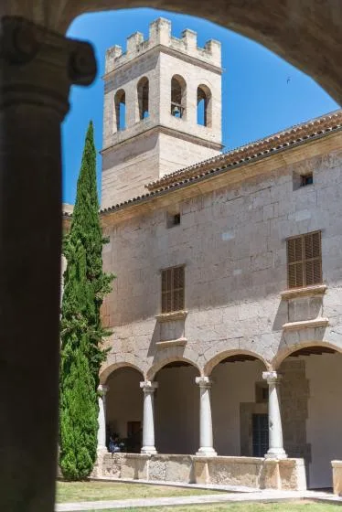 A stone courtyard arcade stands beneath a square bell tower, with a tall cypress nearby.