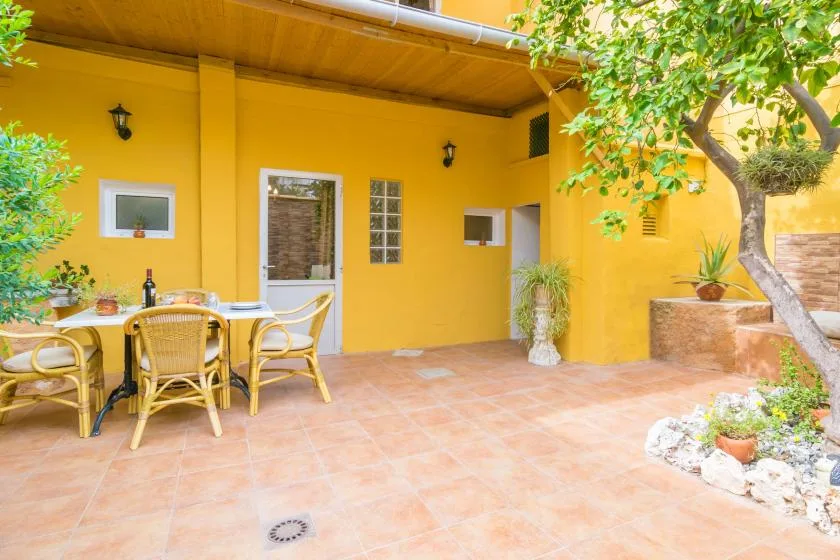 A sunny patio with outdoor seating beside a yellow wall and leafy plants.