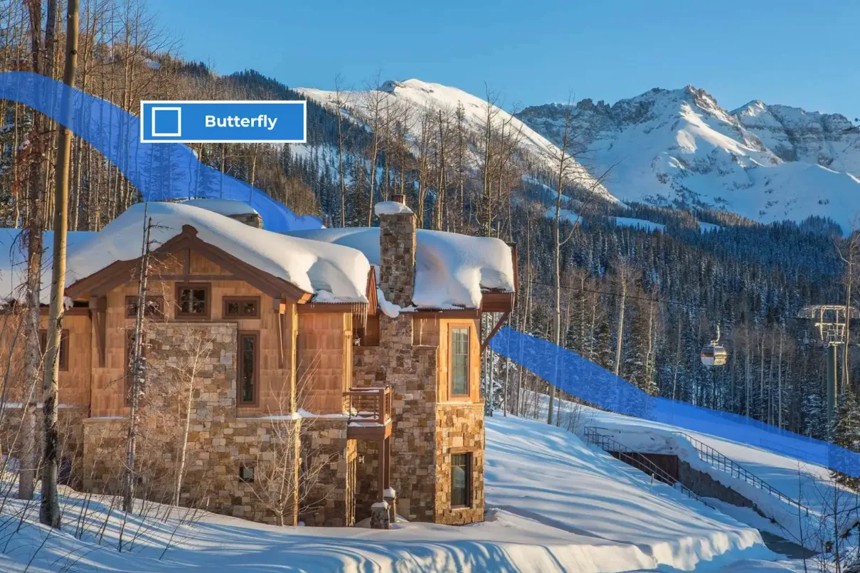 A snow-covered mountain chalet stands among trees, with snowy peaks in the background.