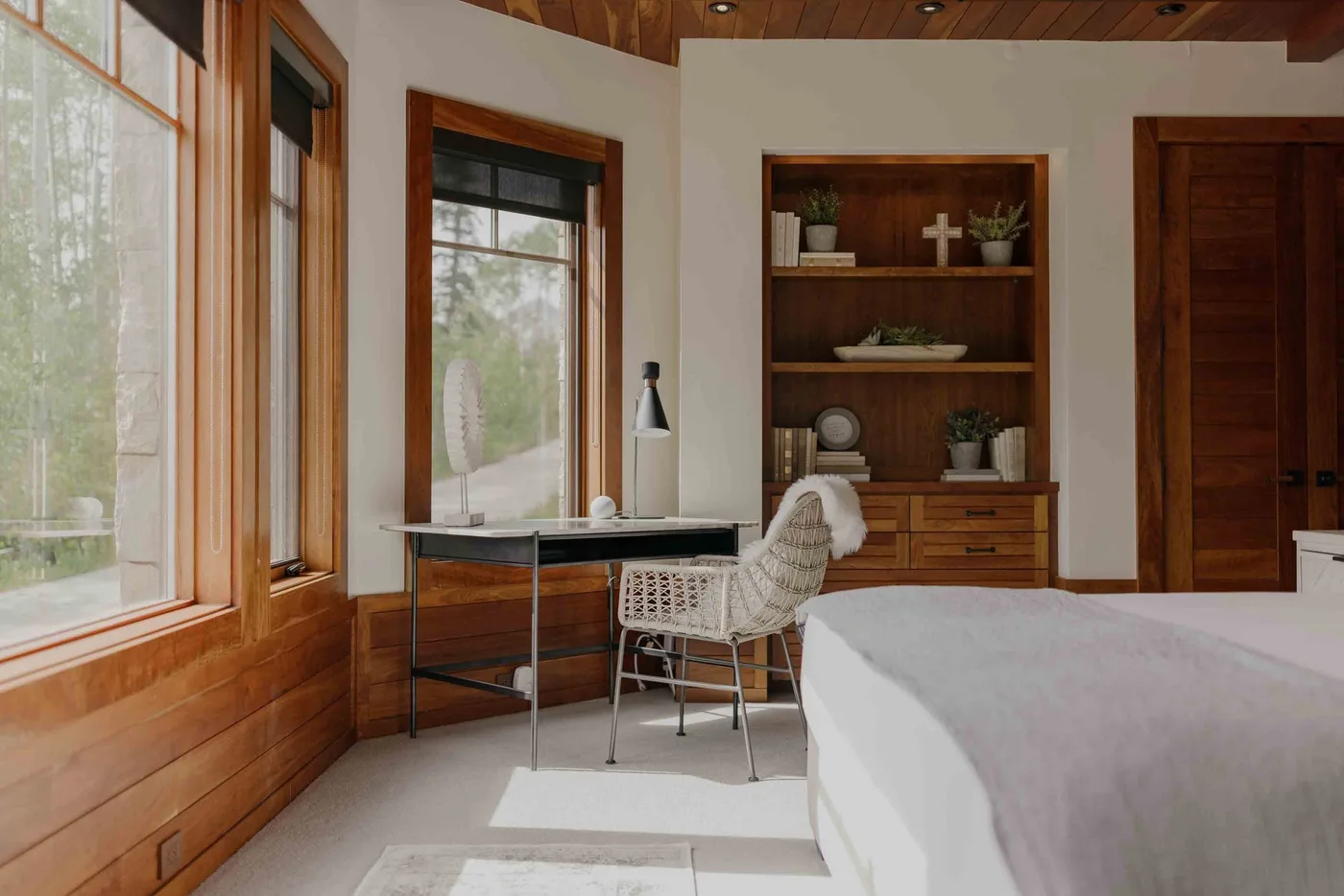 A bedroom with a desk and woven seating beside large wood-framed windows.