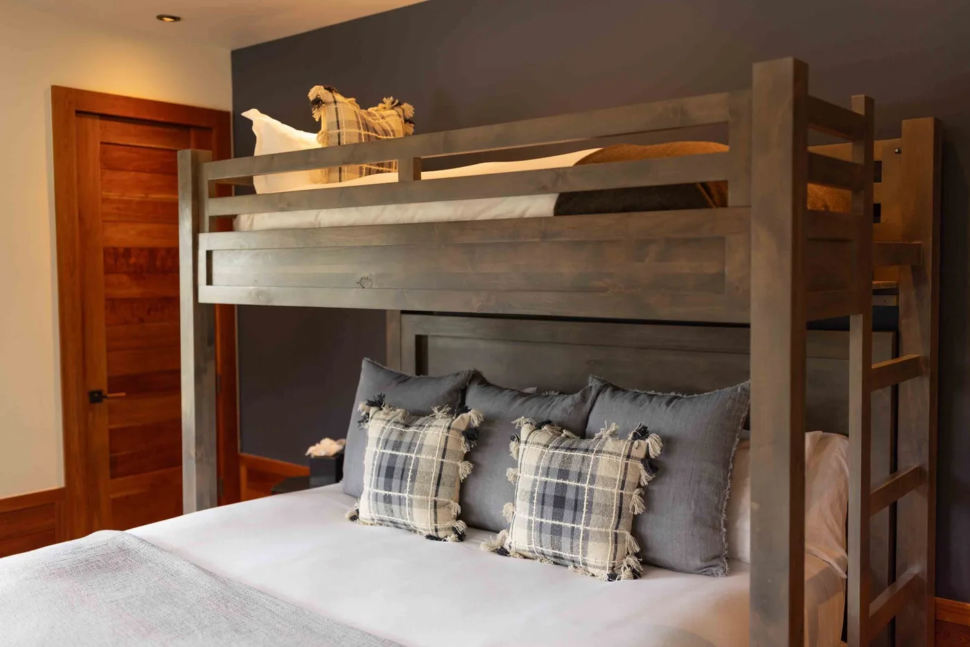 A bedroom with a wooden bunk bed, gray pillows, and plaid cushions.