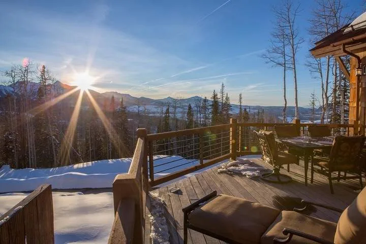 A wooden deck with outdoor seating overlooks a snowy mountain landscape at sunset.