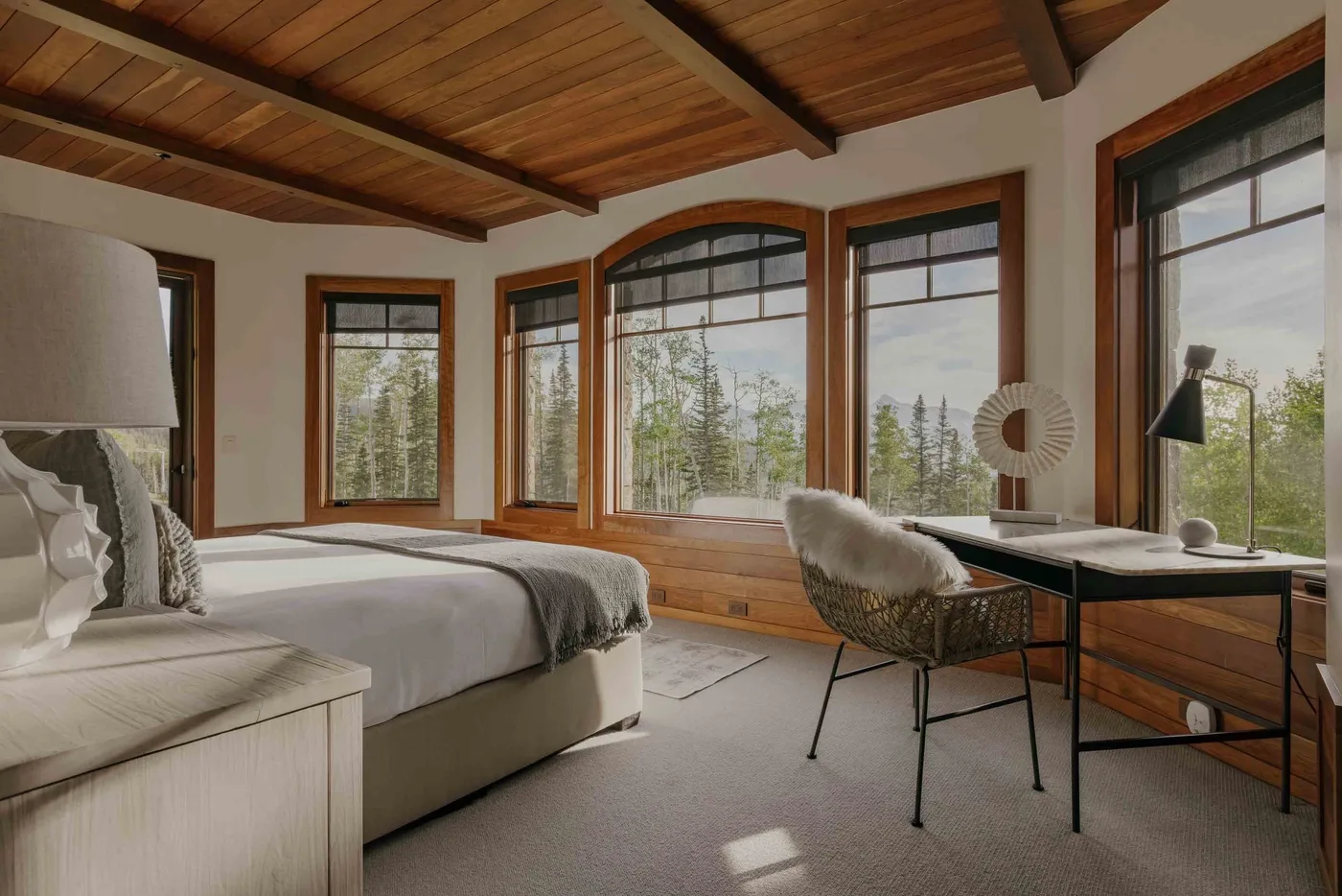 A bedroom with a bed, large windows, and a desk beneath a wood-paneled ceiling.