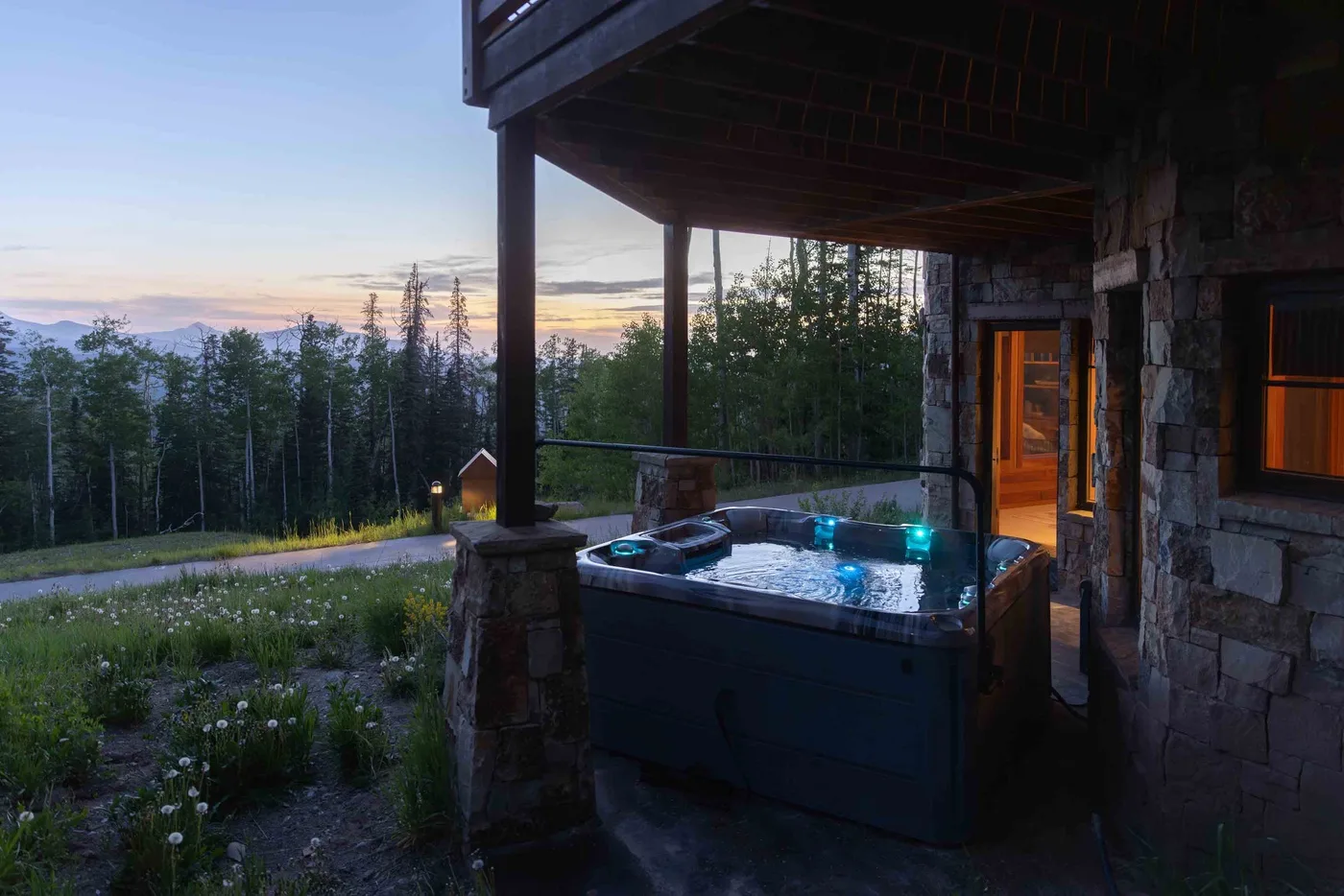 A covered outdoor hot tub sits beside a stone building, overlooking a forest at dusk.