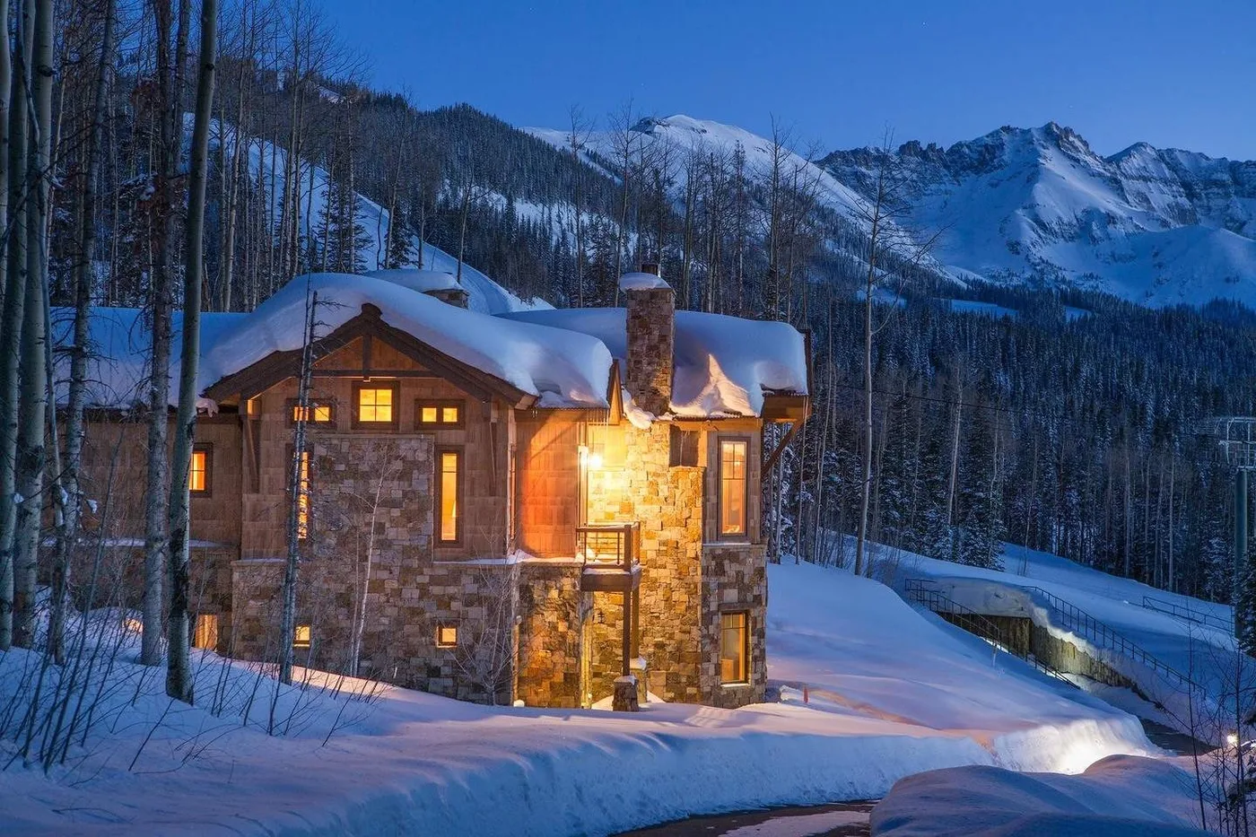 A snow-covered stone house glows among trees, with mountains in the background.
