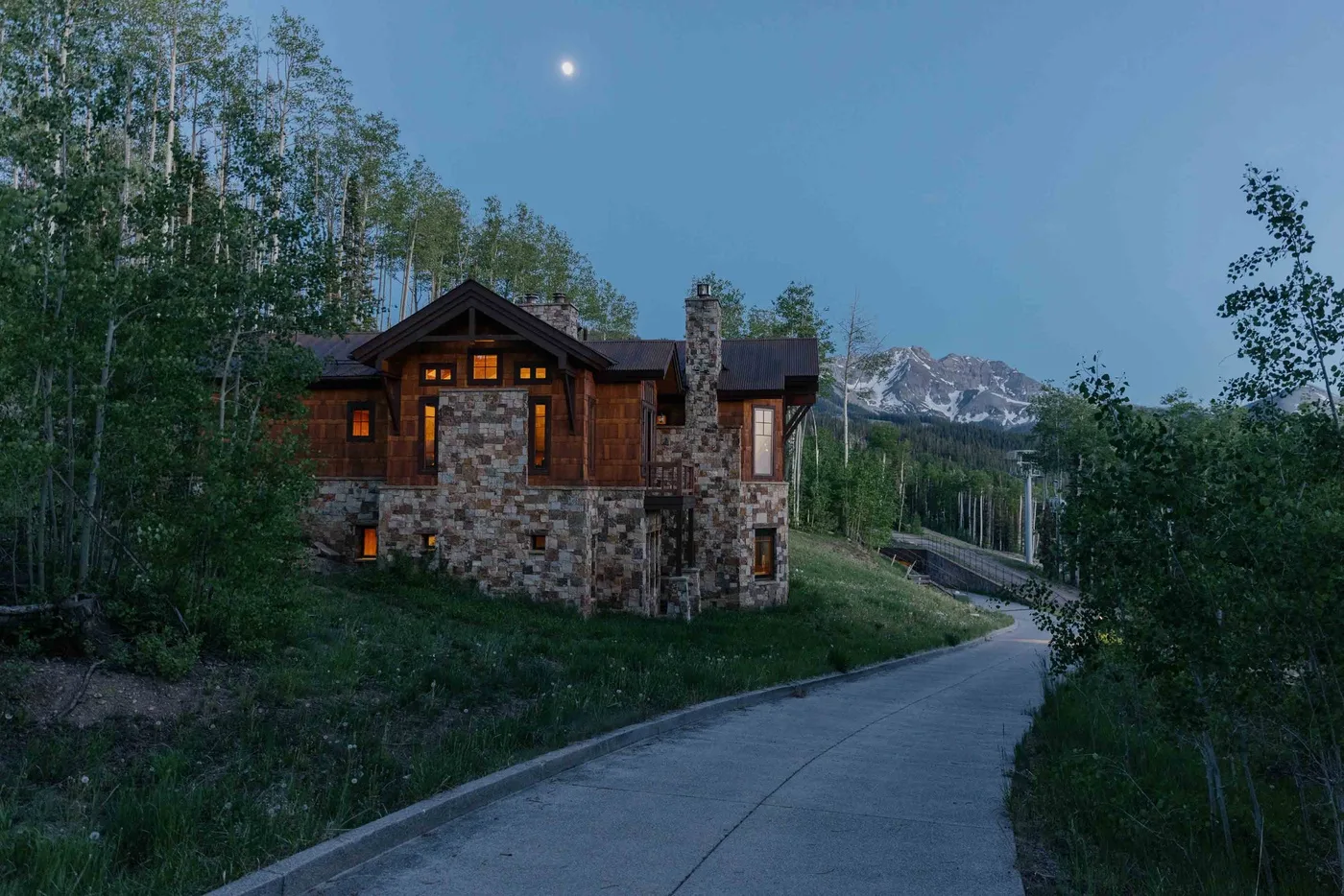 A rustic cabin sits beside a curving paved road, with mountains in the distance.