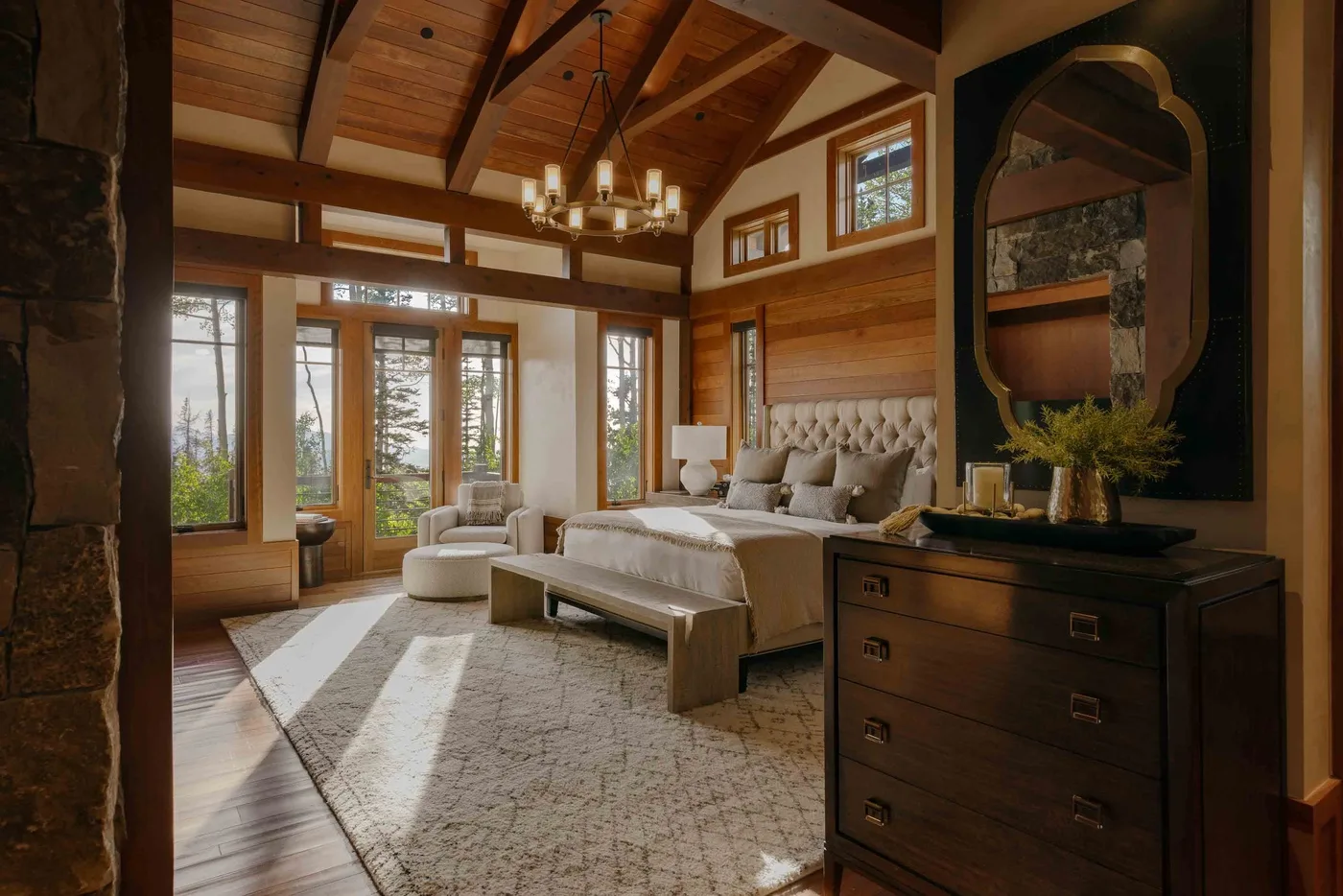A bedroom with a bed, sitting area, and tall windows beneath a wood-beamed ceiling.