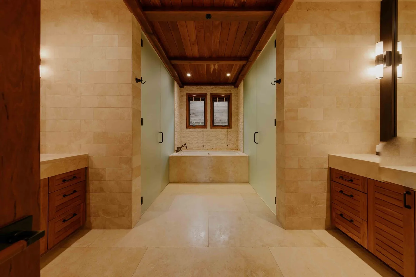 A tiled bathroom with a bathtub beneath two windows and wooden vanities.