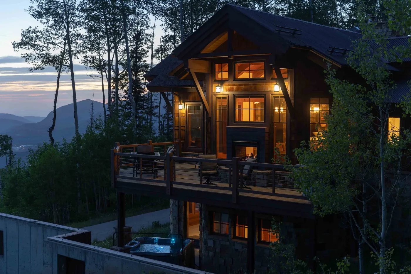 A warmly lit wooden house stands among trees at dusk, with a raised deck.