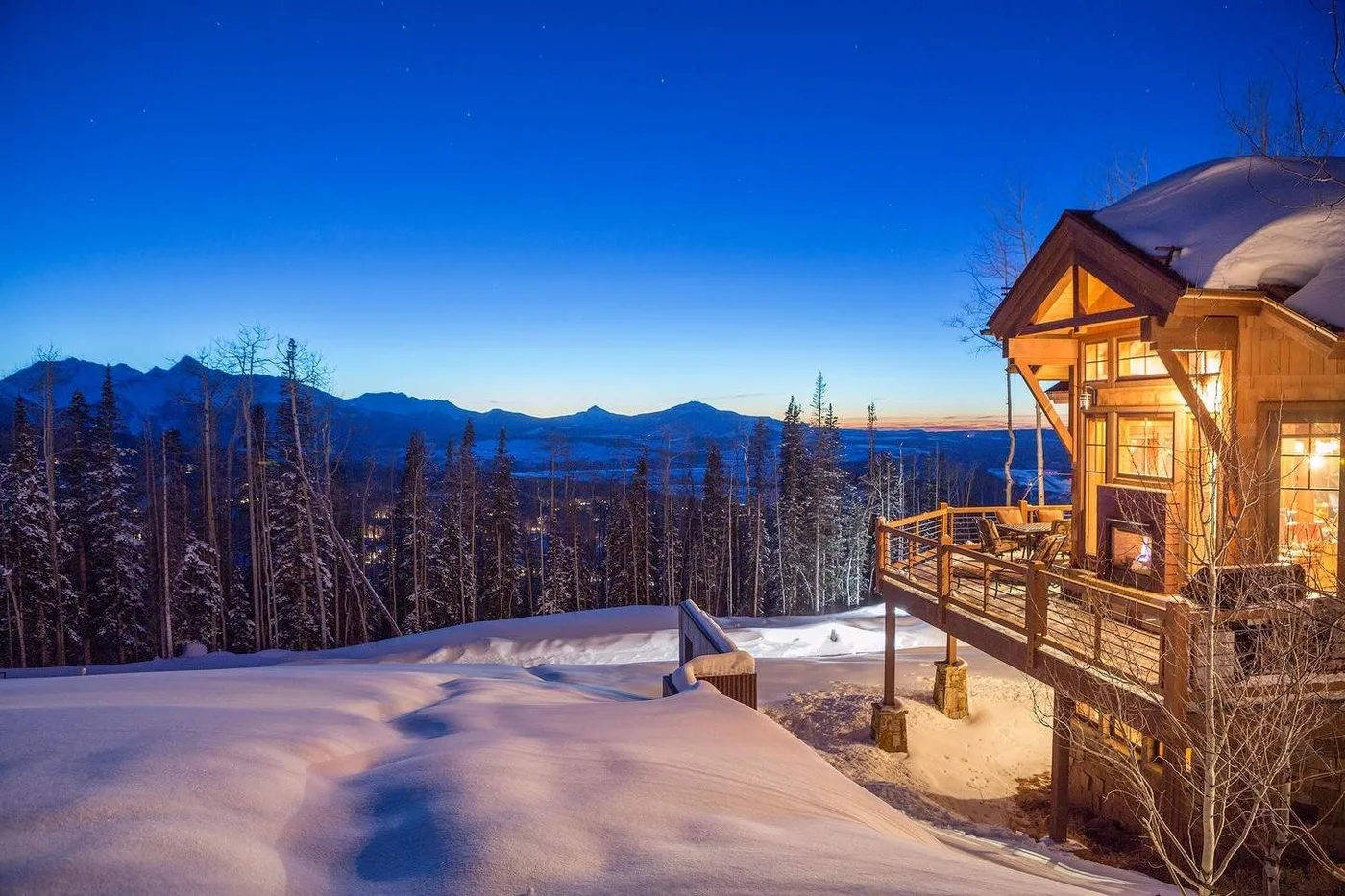 A warmly lit wooden cabin stands beside a snowy forest beneath a mountain skyline.
