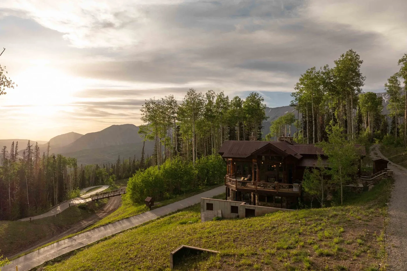 A mountainside cabin sits among trees above a winding road at sunset.