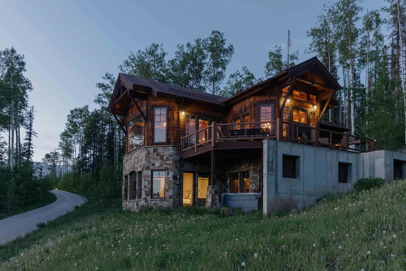 A wood-and-stone house glows with warm lights beside a curving road.
