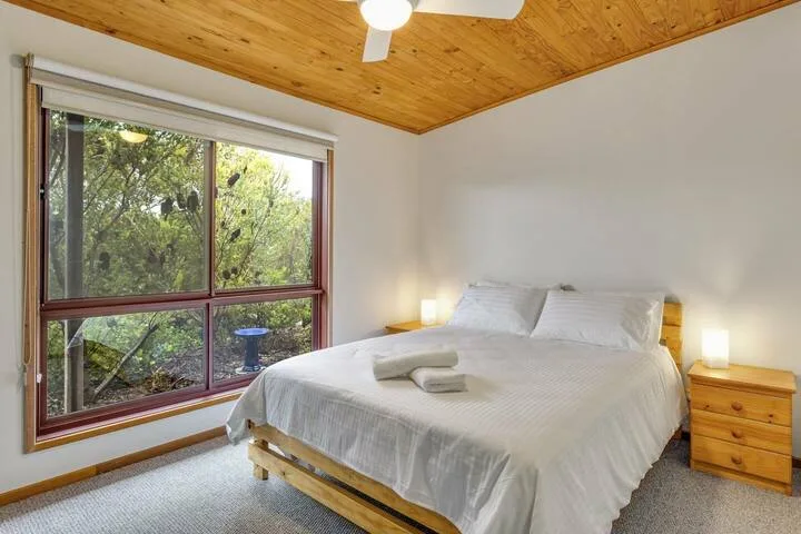 Un dormitorio con una cama junto a una gran ventana con árboles frondosos a la vista.