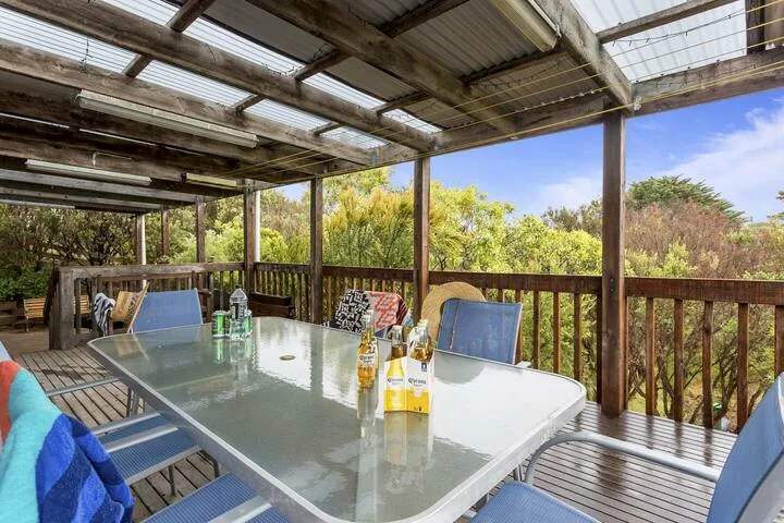 Un comedor exterior cubierto tiene una mesa de vidrio y sillas junto a una barandilla de madera.